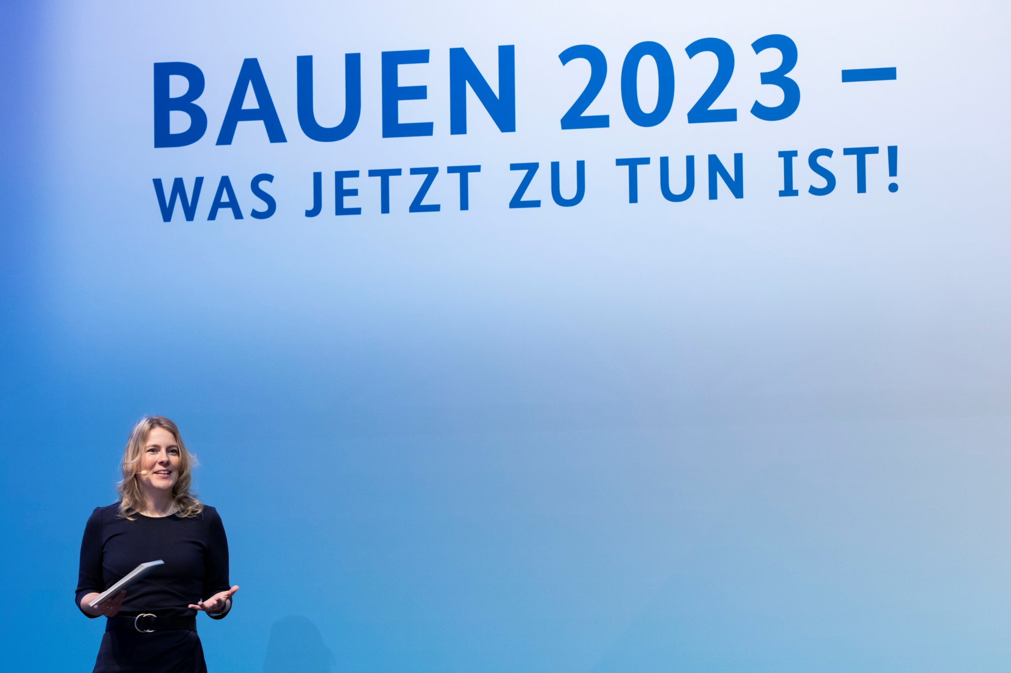 Tina Teucher spricht vor Publikum auf Messe BAU 2023; Text auf Hintergrund: BAUEN 2023 - Was jetzt zu tun ist!