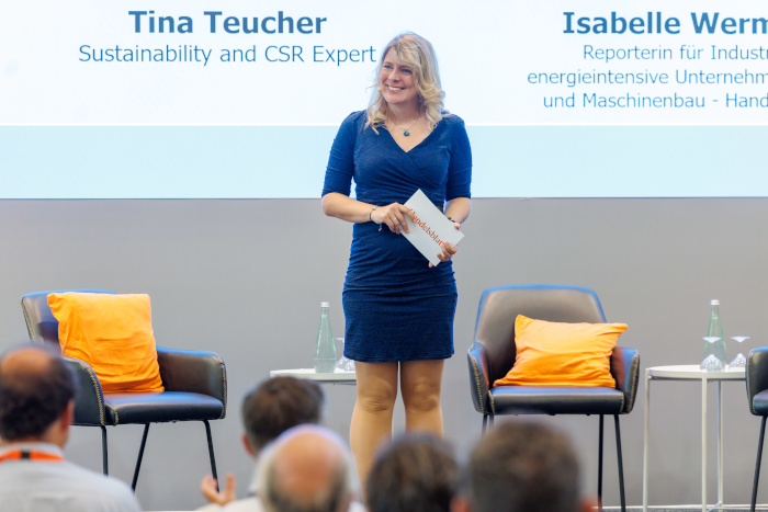 Tina Teucher moderiert auf der Bühne der Handelsblatt Wasserkonferenz 2026, hinter ihr stehen Stühle und die Leinwand ist sichtbar