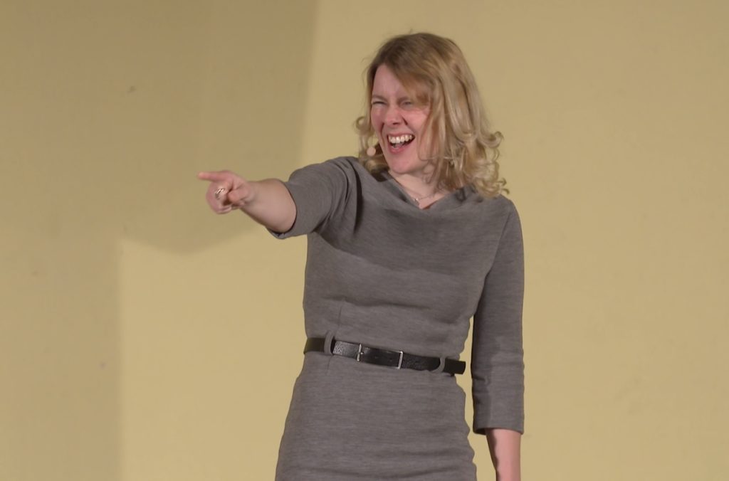 Tina Teucher, eine junge Moderatorin mit blonden Haaren und einem grauen Kleid von Alma und Lovis, zeigt mit ausgestrecktem Arm motivierend an der Kamera vorbei.